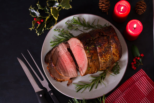 Sliced round roast on a gray plate with rosemary sprigs, next to a carving fork, two lit red candles, holly, pinecones, and a red striped napkin on a dark surface.