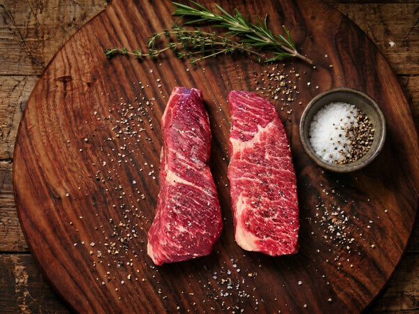 Two raw Denver steaks with salt, pepper and fresh herbs on a wooden board.